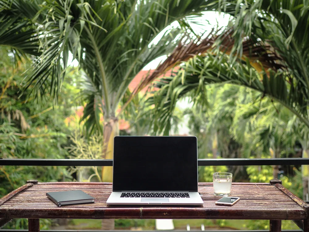 laptop of a remote digital nomad jobs on a wooden bamboo table with notebook, mobile phone and glass in nature with a green tropical background with palm trees
