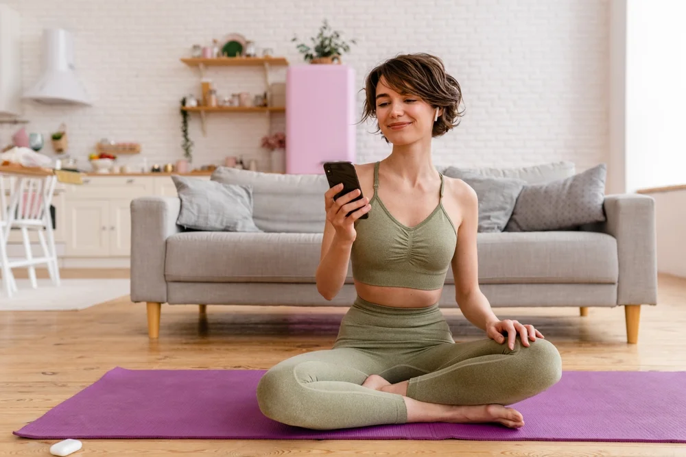 young pretty woman doing yoga at home wearing stylish sport outfit watching exercises on phone, meditation on mat, listening to music in earpods
