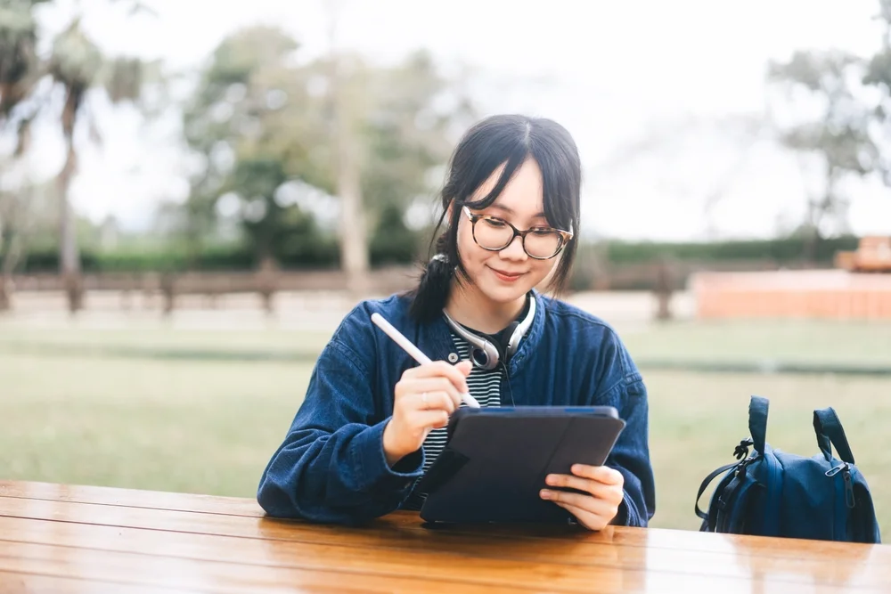 Freelance digital nomad work and study people concept. Young adult asian woman wear eyeglasses and headphone using tablet. Sit on outdoor wooden table. Blue theme colour outfit. Checking expense tracking apps for digital nomads
