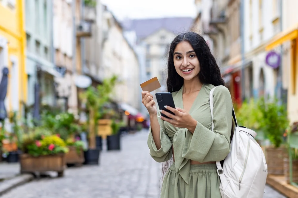 Beautiful young Indian woman using phone and credit card. Standing outside on a city street and smiling at the camera. 
