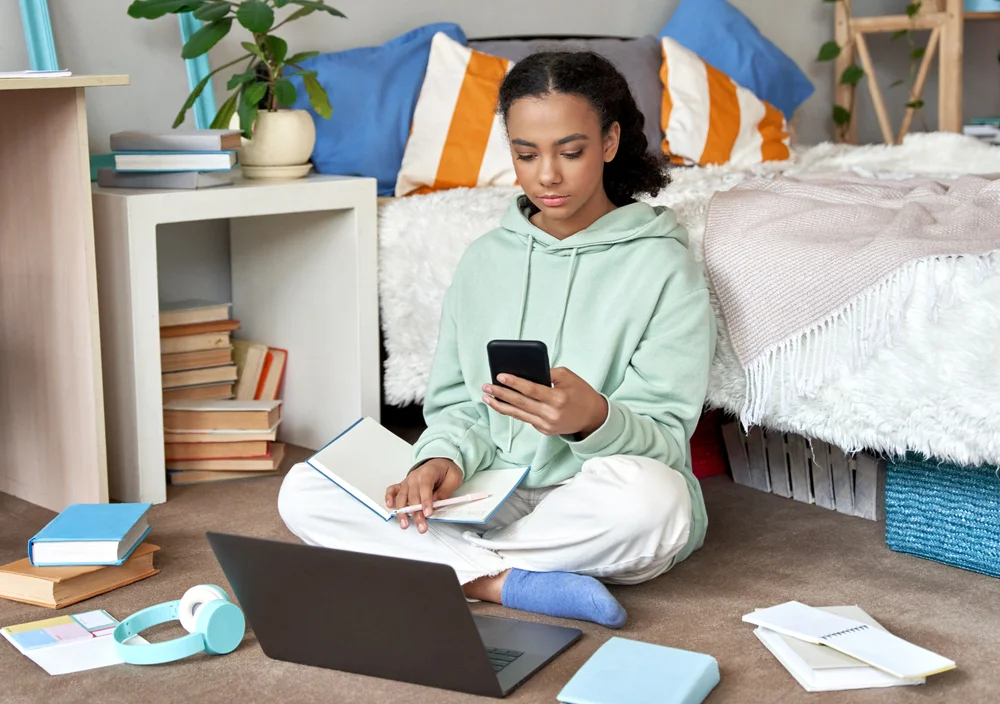 Mixed race teen school girl distant school or college student virtual remote e learning using mobile phone app and laptop in bedroom. Distance education classes, studying online at home concept.
