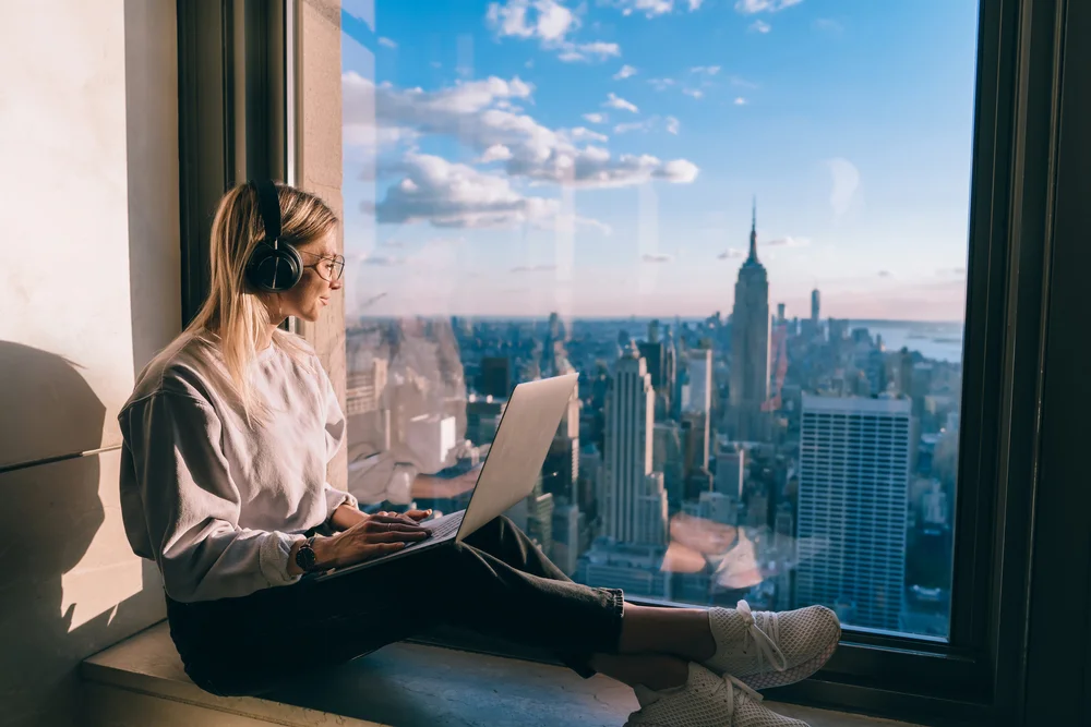 Millennial female freelancer in electronic headphones for noise cancellation listening positive music podcast and looking in panoramic window with breathtaking Manhattan view from high rise
