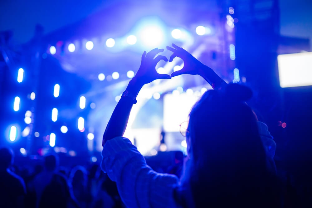 Heart shaped hands at concert, loving the artist and the festival. Music concert with lights and silhouette of people enjoying the concert.
