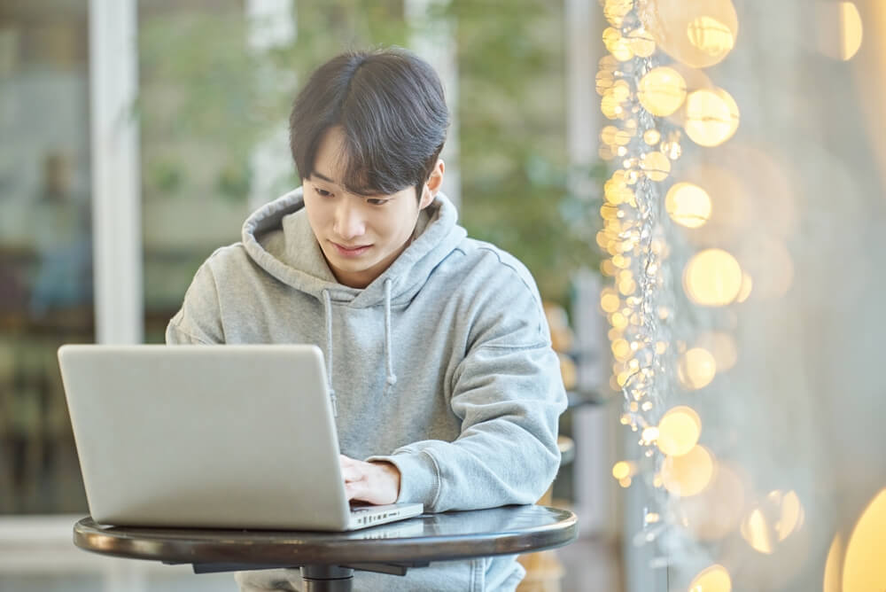 Young male college student model sitting at a cafe table in Asian Korea, listening to a lecture, doing homework or working on a laptop with blurred lights in the background
