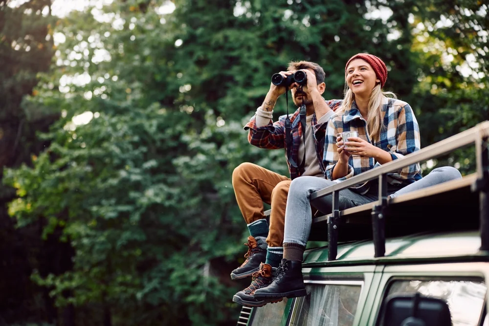 Happy woman van life and her boyfriend using binoculars while sitting on roof of their camper trailer and spending a day in the woods. Copy space.
