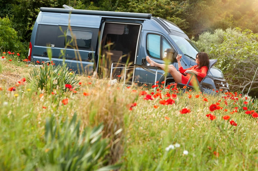 Van life outdoor theme: A dark-haired woman in a red dress sits in an armchair, glass in hand, relaxing in a meadow of blooming red poppies in front of a camper van. Travelling Greece, wanderlust life style.
