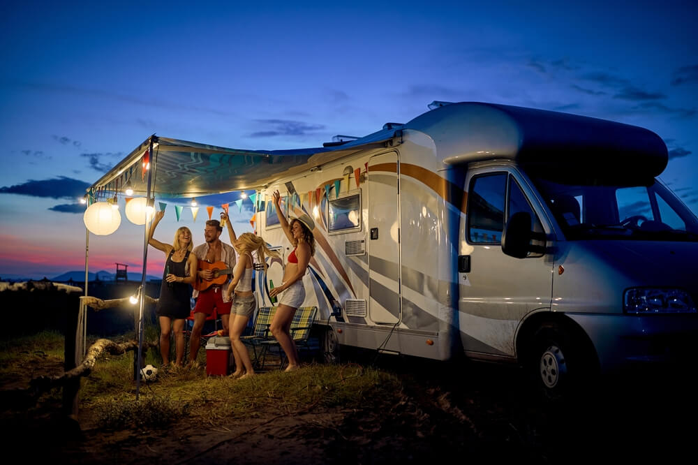 Fun with friends in camp life community, in front of rv. Young group of people dancing, playing guitar, and celebrating. Summertime togetherness. Travel, weekend, lifestyle concept.
