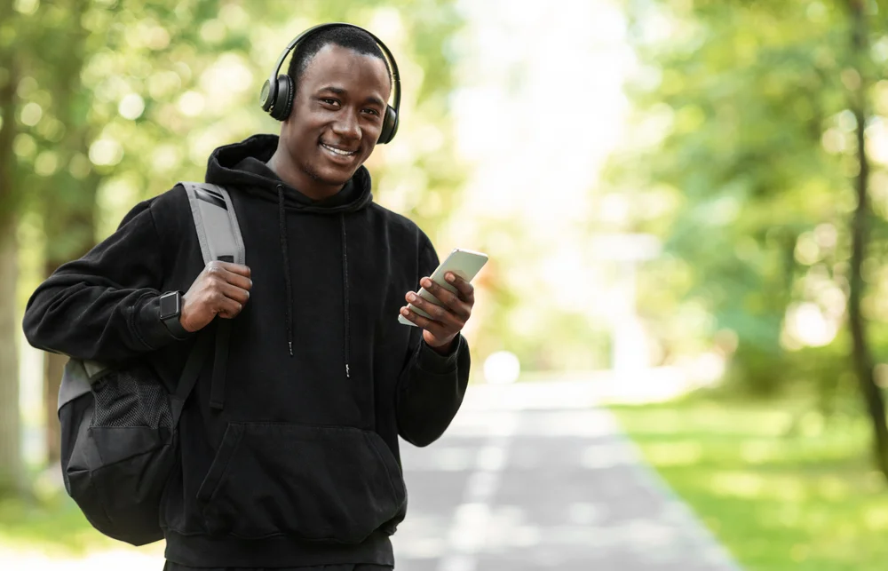 Black guy in sport clothes and backpack using phone, going home after training outdoors, free space