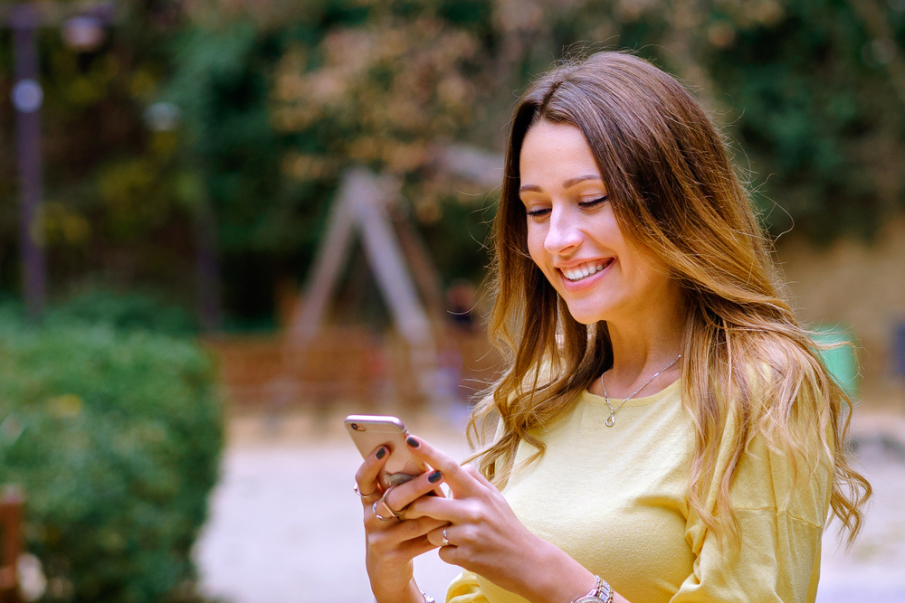 Beautiful young woman in trendy outfit smiling and browsing smartphone while standing on blurred background of park