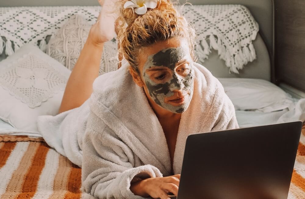 Pretty adult woman with skin beauty cream on face work on laptop computer laying in campervan essentials bedroom. Travel lifestyle female people with online job. Modern lady and healthy treatment
