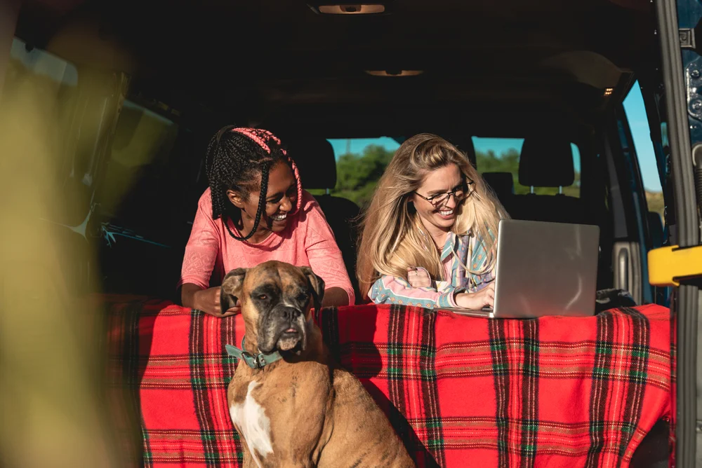 Multiracial people with dog traveling together retro mini van transport - Digital nomad concept working at laptop computer - Focus on african girl face.