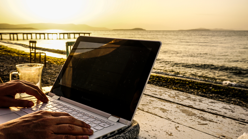 Freelance female hands working with laptop on wooden table on beach at sunset. Woman sitting on chair and using computer near blue sea shore at sunny day. Digital nomadism towards yellow sky & horizon
