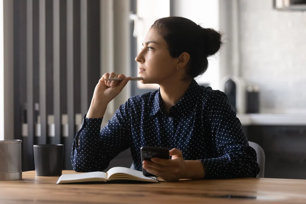 Pensive young hindu female study by desk using mobile internet distracted from setting goals create new idea. Thoughtful mixed race woman looking aside of phone screen pondering planning future work
