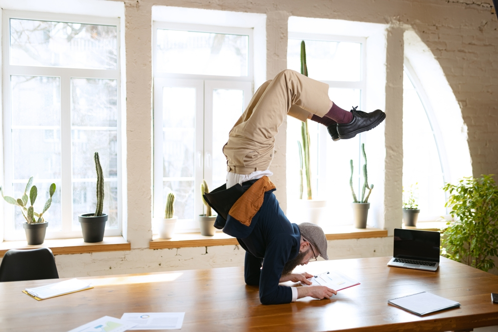 Flexibility. Man practicing yoga. Young bearded office clerk having fun, doing yoga on wooden table in modern office at work time with gadgets. Concept of business, healthy lifestyle, sport, hobby

