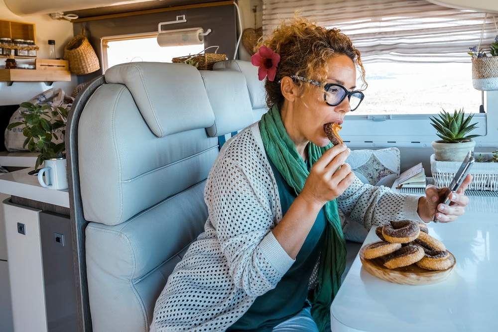 Bored woman eating a lot of donuts sitting at the table inside a camper van during travel lifestyle. Female people enjoy vacation in motorhome having relax and using mobile phone and connection
