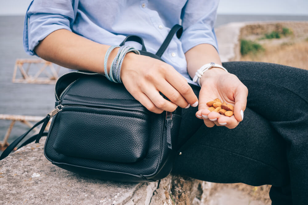 Young woman having a healthy snack, while sitting on a parapet near the sea. Female's hand holding a handful of almonds next to an open bag, close-up.
