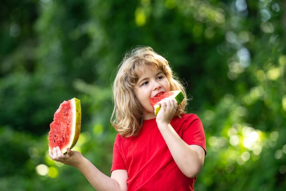 Happy Kid with watermelon. Funny kid eating watermelon outdoors in summer park. Child, baby, healthy food. Child eating watermelon in the garden. Kids eat fruit outdoors. Kid enjoying watermelon.
