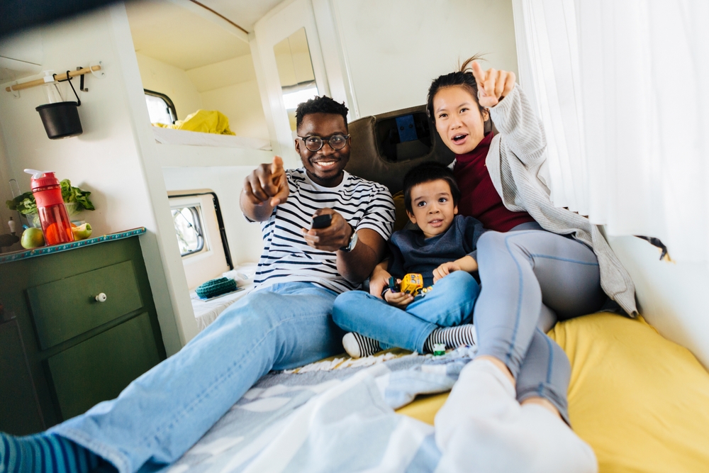 A fun family is lying in the bed in their motor home and watching movies and cartoons on TV stream. The mother is pointing at the TV stream while the father holding a remote control.
