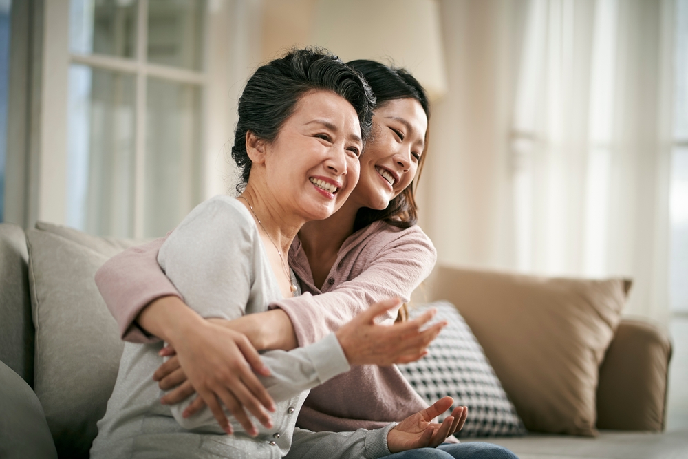 loving Japan adult daughter sitting on couch at home hugging senior mother happy and smiling
