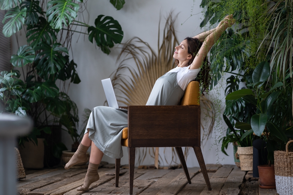 Balancing Work and Personal Life. Happy female freelancer with closed eyes relaxing while working remotely in home garden full of exotic plants. Young woman resting during remote work at urban jungle home office
