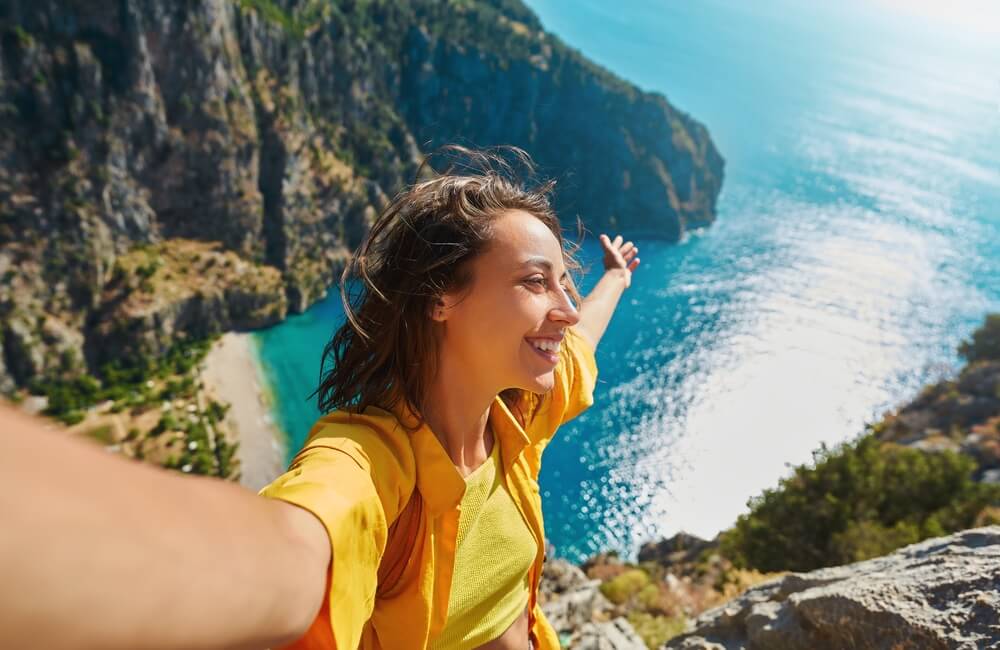 Positive tourist woman taking picture outdoors for memories, making selfie on top of cliff with valley mountains view, sharing travel adventure journey
