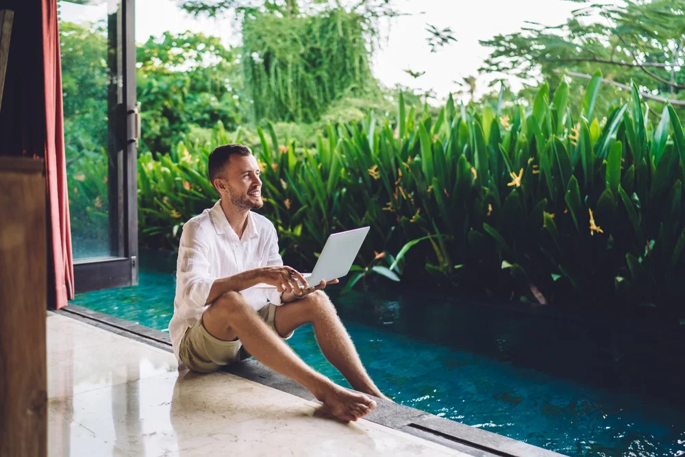 Successful copywriter with modern laptop device enjoying time at pool terrace during summer vacations at Bali, happy male digital nomad with netbook computer smiling outdoors - freelance lifestyle
