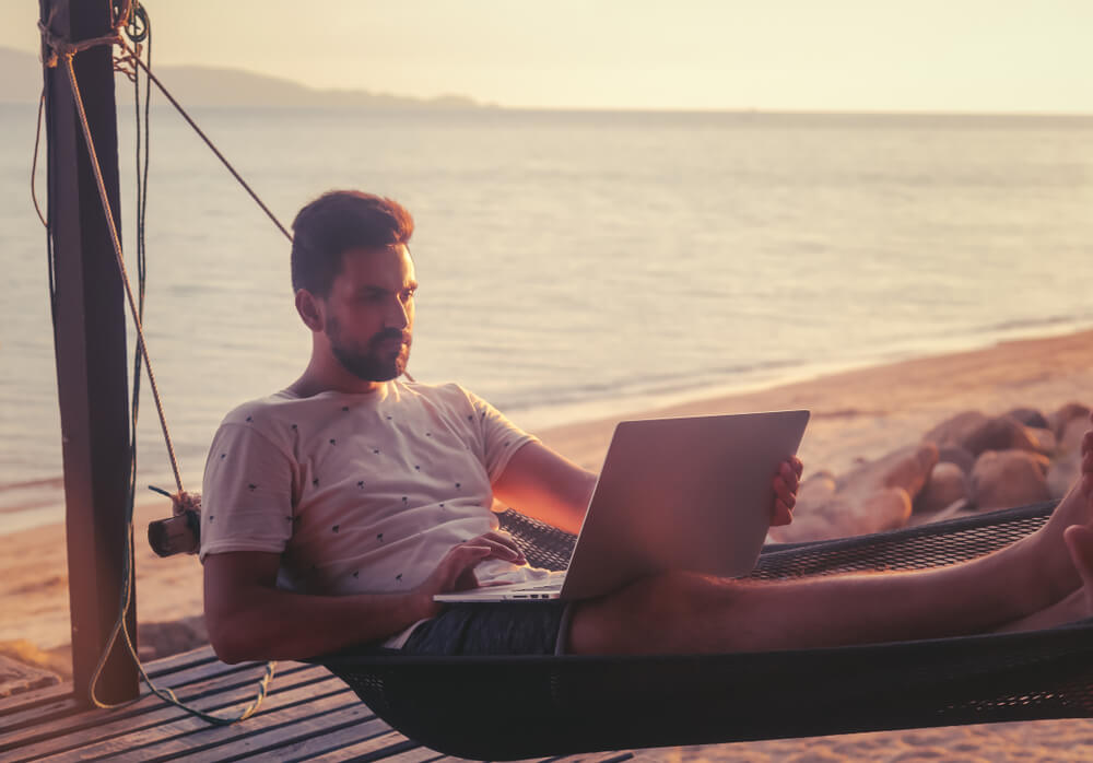 Young attractive guy a man with a beard in a hammock with a laptop on the background of the sea and sunset, distant work, freelancer, blogger, vacation and travel
