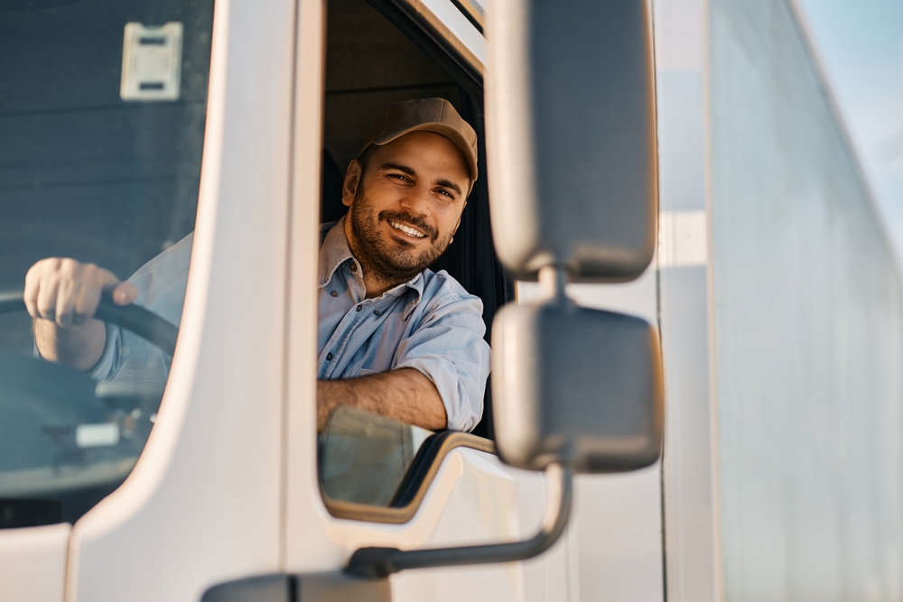 Happy professional truck driver driving his truck and looking at camera by looking for the best wifi for truckers. Copy space.
