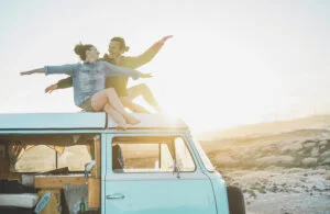 Happy couple sitting on top of minivan roof at sunset - Young people having fun on summer vacation traveling around the world - Travel, love, van lifestyle and freedom concept - Focus on bodies
