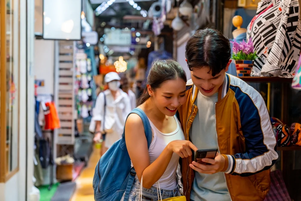 Asian couple enjoy and fun outdoor lifestyle shopping at street market on summer holiday vacation. Happy man and woman couple using mobile phone together while walking and shopping at weekend market.