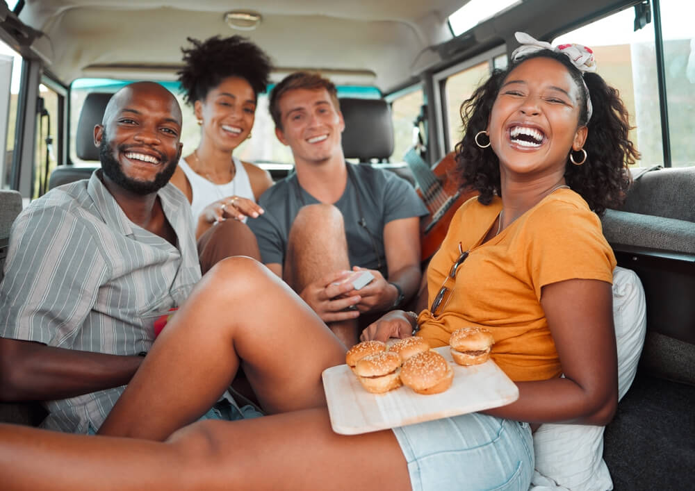 Food, travel and friends eating on a road trip, happy, relax and laughing while bonding in a car together. Fast food, diversity and face portrait of smiling people enjoying freedom and adventure trip
