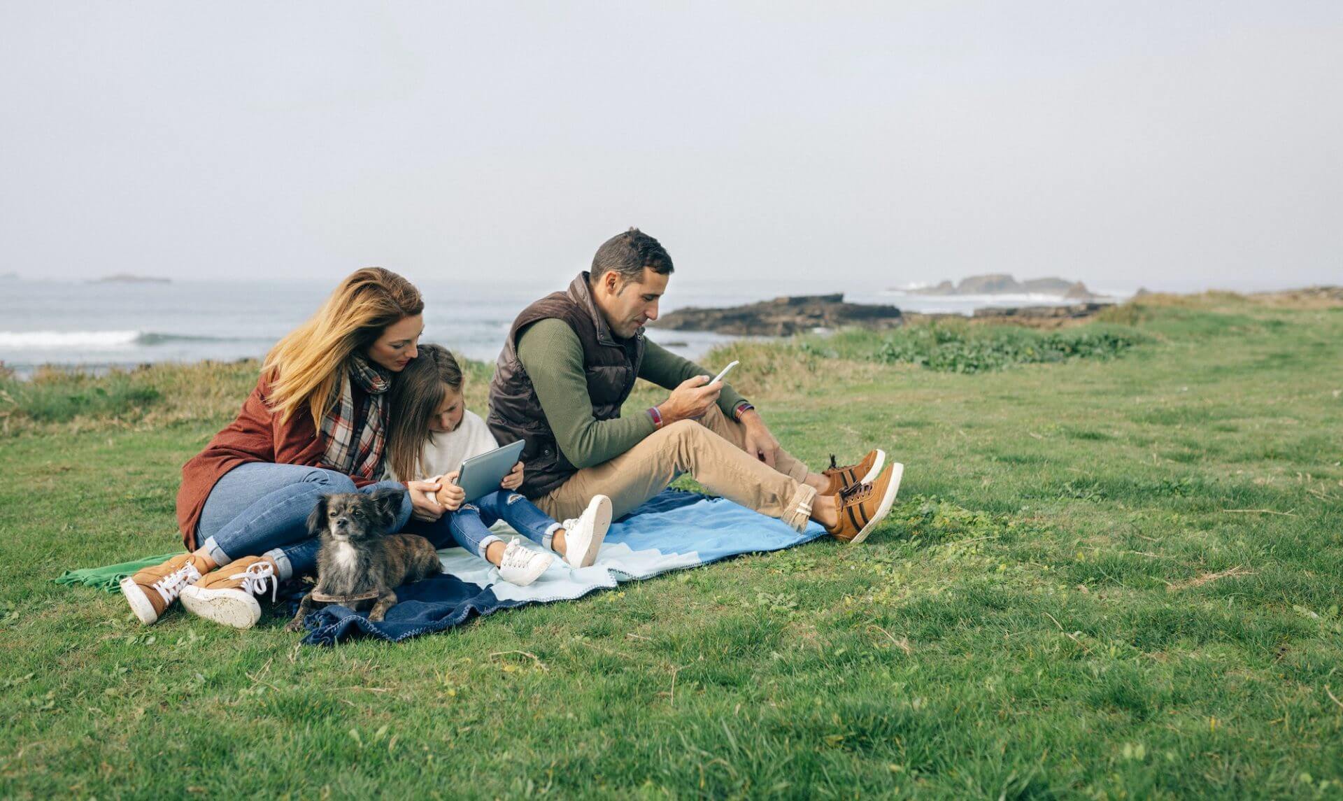 Family with dog sitting on blanket at the coast using wireless devices