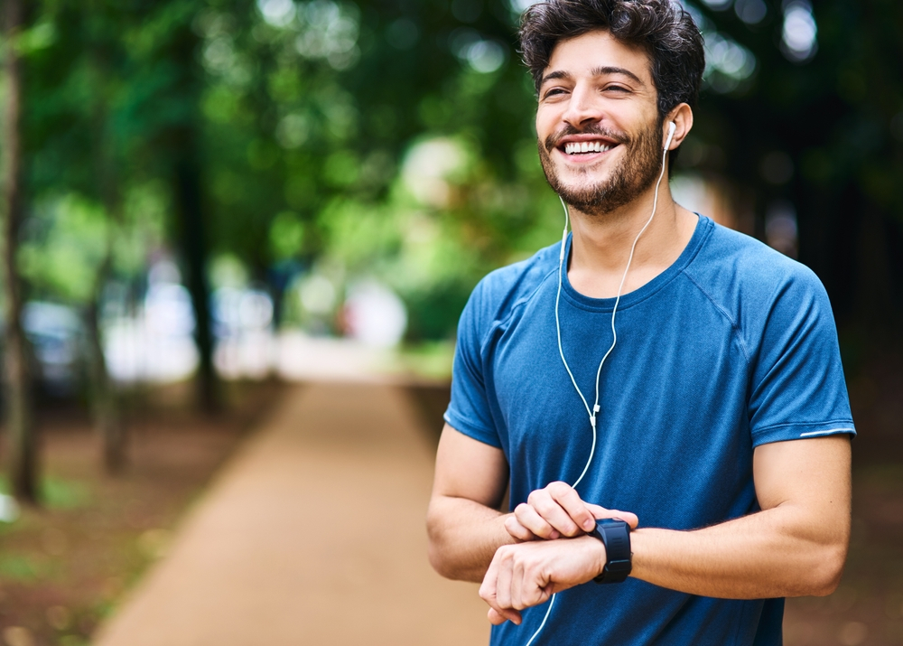 Fitness, music and man with a watch for time, running progress and heart results in a park. Smile, thinking and a male athlete listening to a podcast or audio while checking notification on a device
