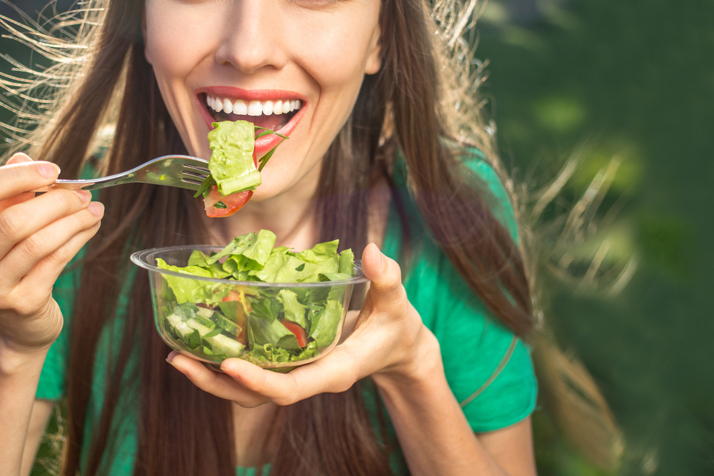 Woman eating healthy salad from plastic container over green grass with flying hair on a sunny spring day. backlight
