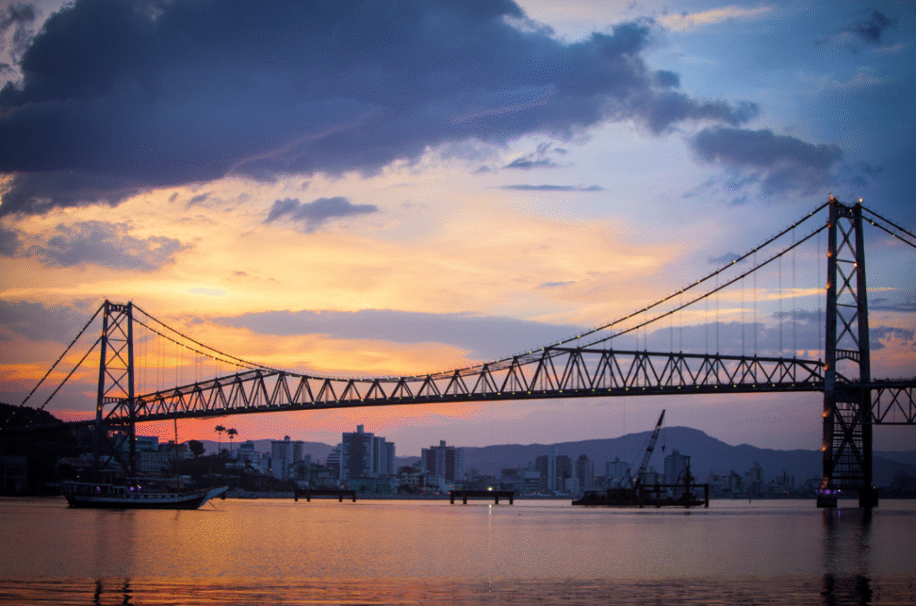 Hercílio Luz Bridge at sunset in Florianópolis, Brazil, with colorful sky reflecting on the water and city skyline in the background.
