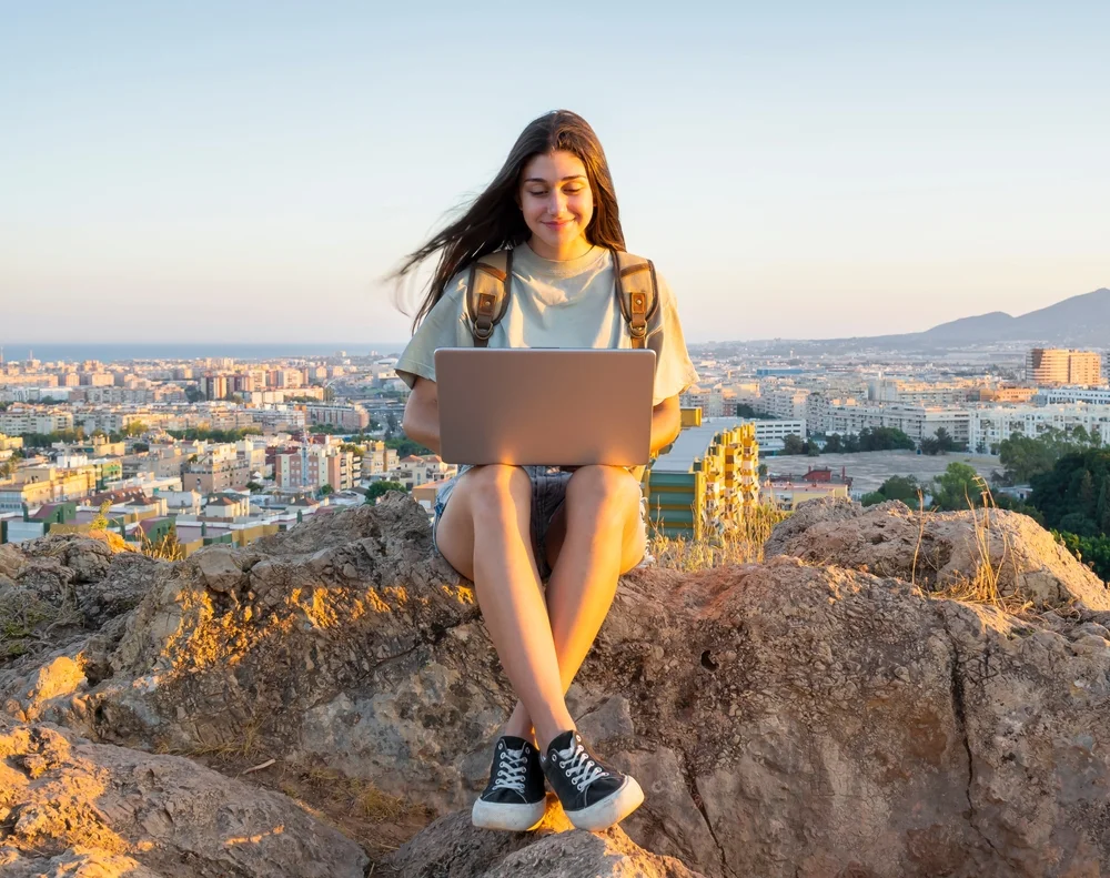Young woman with a backpack, using a laptop to make a video call in the middle of a field. Digital nomad. In the background Malaga at sunset.