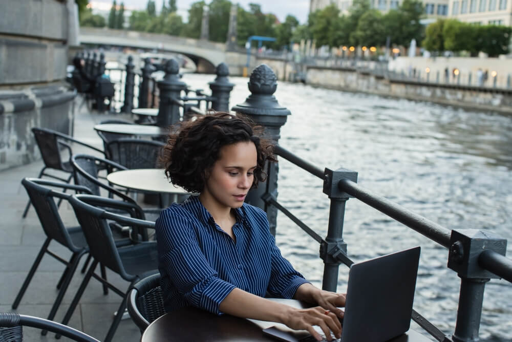 curly young woman using laptop while sitting on summer terrace near river in berlin
