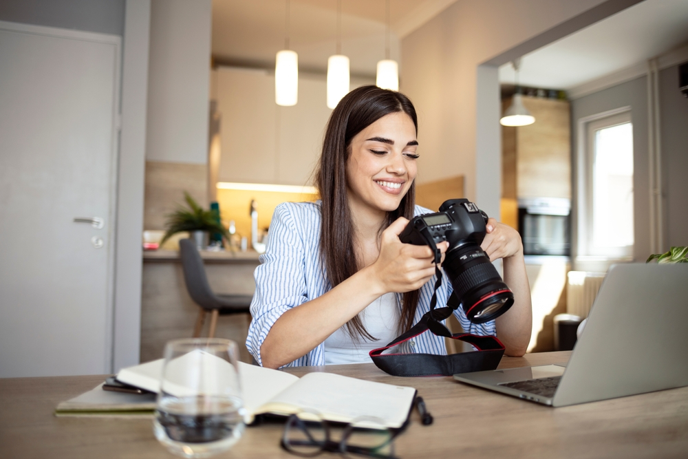 Happy young photographer holding a dslr camera in her home office. Female photographer smiling cheerfully while working at her desk. Creative female freelancer working on a new project. She has a successful remote career
