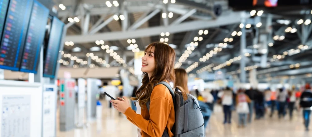 Young asian woman in international airport, using mobile smartphone and checking flight at the flight information board
