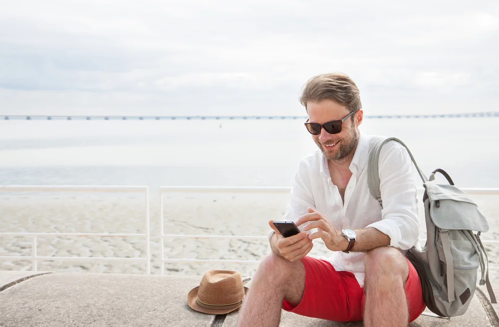 Happy smiling forty years old caucasian tourist man looking at mobile phone outdoor. Beach and sea as background - summer holiday traveling.
