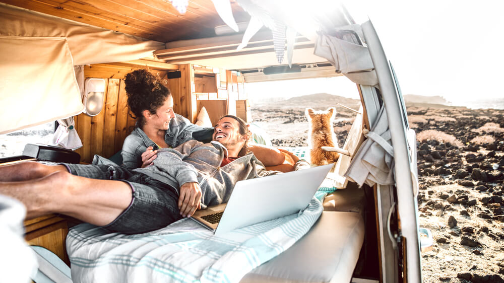Hipster man and woman with dog traveling together on mini van life transport - Freelance nomad concept with hippie people on minivan romantic trip working at laptop pc in relax moment - Warm retro filter
