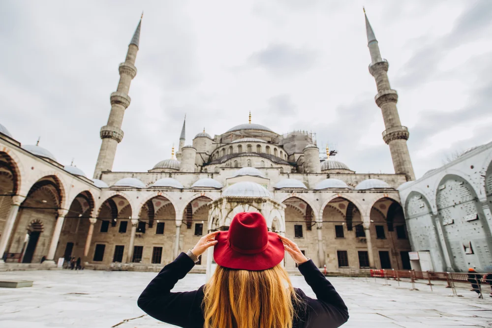 Young woman traveler in hat and black coat looking at the blue mosque in Istanbul, Turkey. A girl walks through winter Istanbul. Photo of travel blonde on the background of a mosque on autumn day.
